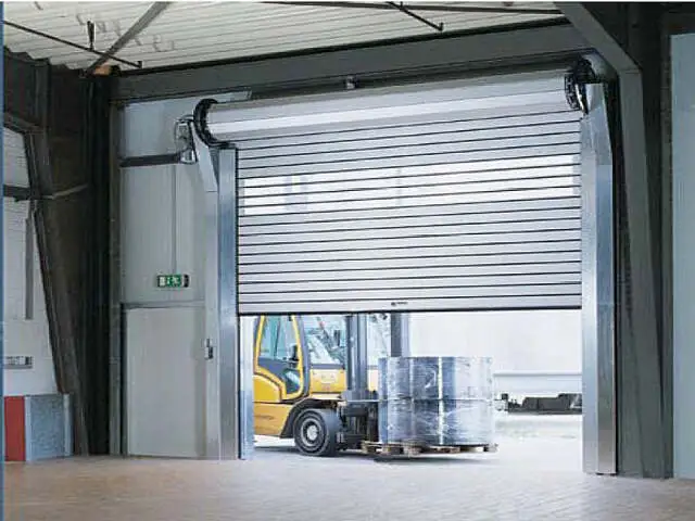 Una gran puerta corrediza industrial está parcialmente abierta, permitiendo el paso de un montacargas que lleva múltiples pilas de mercancías envueltas. El interior está bien iluminado y se ve una señal de salida.